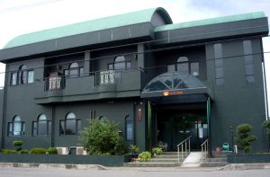 2階建ての介護施設の正面外観。緑色の外壁に複数の窓があり、入口上部には装飾的なアーチ。